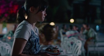 Movie still from “Aftersun” (2022), directed by Charlotte Wells – A young girl holding a plate of food in her hands; Medium shot, Over the shoulder angle