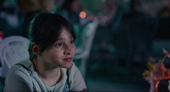 Movie still from “Aftersun” (2022), directed by Charlotte Wells – A young girl sitting in front of a group of people; Close Up shot, Over the shoulder angle