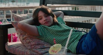 Movie still from “Aftersun” (2022), directed by Charlotte Wells – A young girl sitting in a chair with a drink in front of her; Medium shot, High angle