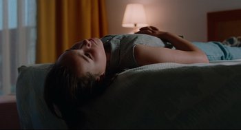 Movie still from “Aftersun” (2022), directed by Charlotte Wells – A young girl laying on a bed in a room with a lamp; Medium shot, Low angle