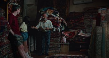 Movie still from “Aftersun” (2022), directed by Charlotte Wells – An older man standing next to a young girl in front of a pile of rugs; Wide shot, High angle