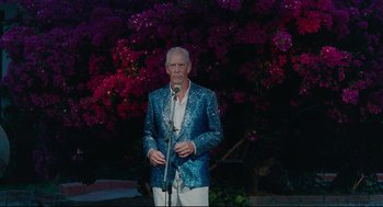 Movie still from “Aftersun” (2022), directed by Charlotte Wells – An older man in a blue jacket is holding a microphone; Medium shot, Low angle