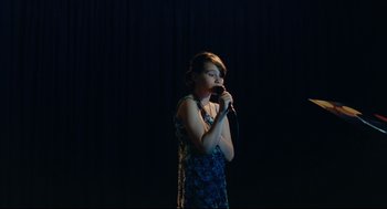 Movie still from “Aftersun” (2022), directed by Charlotte Wells – A woman in a blue dress holding a microphone in her hands; Medium shot, High angle