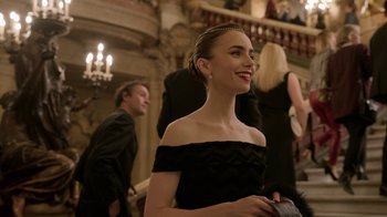 Movie still from “Emily in Paris” (2020), created by Darren Star – A woman in a black dress smiling at a formal event; Medium shot, Over the shoulder angle