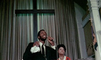 Movie still from “Ganja & Hess” (1973), directed by Bill Gunn – A man and a woman singing into microphones in front of a cross; Medium shot, Low angle