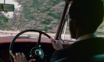Movie still from “Ganja & Hess” (1973), directed by Bill Gunn – A man driving a car while holding a cell phone; Close Up shot, Over the shoulder angle