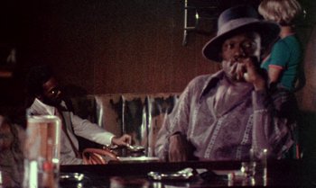 Movie still from “Ganja & Hess” (1973), directed by Bill Gunn – A man sitting at a table smoking a cigarette; Medium shot, Low angle