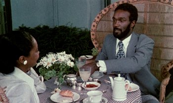 Movie still from “Ganja & Hess” (1973), directed by Bill Gunn – Two people sitting at a table with food and drinks; Medium shot, Over the shoulder angle