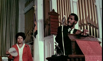 Movie still from “Ganja & Hess” (1973), directed by Bill Gunn – A man speaking at a podium in front of an audience; Medium shot, Low angle