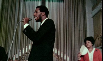 Movie still from “Ganja & Hess” (1973), directed by Bill Gunn – A man in a black suit singing into a microphone; Close Up shot, Low angle