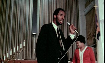 Movie still from “Ganja & Hess” (1973), directed by Bill Gunn – A man in a black suit is speaking into a microphone; Close Up shot, Low angle