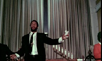 Movie still from “Ganja & Hess” (1973), directed by Bill Gunn – A man in a suit holding a wii controller in front of a row of lit candles; Medium shot, Low angle