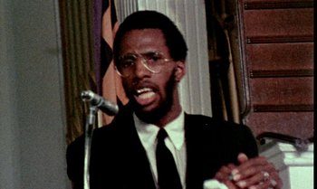 Movie still from “Ganja & Hess” (1973), directed by Bill Gunn – A man in a black suit and white tie is speaking into a microphone; Close Up shot, Low angle