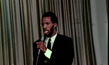 Movie still from “Ganja & Hess” (1973), directed by Bill Gunn – A man wearing a suit and tie holding a microphone; Close Up shot, Low angle