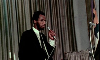 Movie still from “Ganja & Hess” (1973), directed by Bill Gunn – A man in a suit and tie holding a microphone; Close Up shot, Low angle