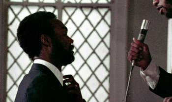 Movie still from “Ganja & Hess” (1973), directed by Bill Gunn – A man in a suit and tie holding a microphone; Close Up shot, Over the shoulder angle