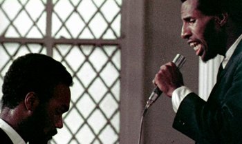 Movie still from “Ganja & Hess” (1973), directed by Bill Gunn – A man holding a microphone in front of another man in a suit; Close Up shot, Low angle