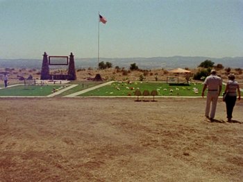 Movie still from “Gates of Heaven” (1978), directed by Errol Morris – A man standing in a field near a flag pole; Extreme Wide shot, High angle