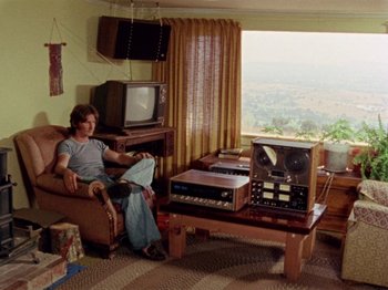 Movie still from “Gates of Heaven” (1978), directed by Errol Morris – A man sitting in a chair in front of a television set; Wide shot, High angle