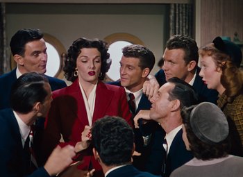 Movie still from “Gentlemen Prefer Blondes” (1953), directed by Howard Hawks – A group of people gathered around a woman in a red suit; Medium shot, High angle