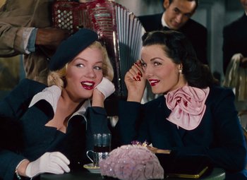 Movie still from “Gentlemen Prefer Blondes” (1953), directed by Howard Hawks – Two beautiful women sitting at a table with a cake; Medium shot, Low angle