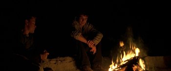 Movie still from “Gerry” (2002), directed by Gus Van Sant – A man sitting in front of a fire at night; Medium shot, Low angle