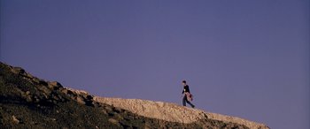 Movie still from “Gerry” (2002), directed by Gus Van Sant – A man walking on top of a hill holding a skateboard; Extreme Wide shot, Low angle