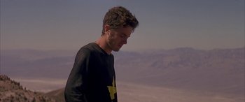 Movie still from “Gerry” (2002), directed by Gus Van Sant – A young man with a black t - shirt is looking down at the ground; Medium shot, Low angle