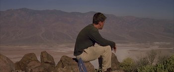 Movie still from “Gerry” (2002), directed by Gus Van Sant – A man sitting on top of a rock on top of a mountain; Wide shot, Low angle