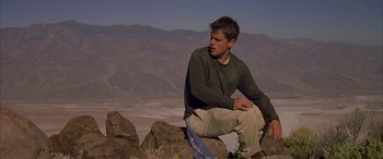 Movie still from “Gerry” (2002), directed by Gus Van Sant – A man sitting on top of a rock in the middle of the desert; Medium shot, Low angle