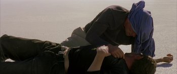 Movie still from “Gerry” (2002), directed by Gus Van Sant – A man is lying on the ground with another man on his back; Medium shot, Over the shoulder angle