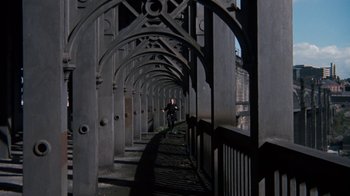 Movie still from “Get Carter” (1971), directed by Mike Hodges – A person walking on a bridge with a fence in front of them; Extreme Wide shot, High angle