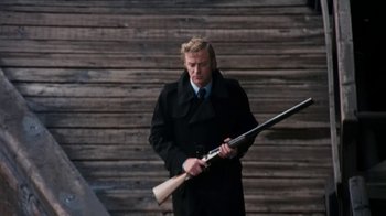 Movie still from “Get Carter” (1971), directed by Mike Hodges – A man holding a rifle in front of a wooden wall; Medium shot, Low angle