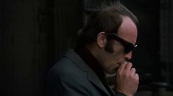 Movie still from “Get Carter” (1971), directed by Mike Hodges – A man in a black suit and sunglasses is holding his hand to his face; Close Up shot, Low angle