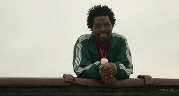 Movie still from “Ghost Dog: The Way of the Samurai” (1999), directed by Jim Jarmusch – A man smiling while sitting on top of a brick wall; Medium shot, Low angle