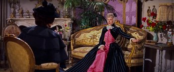 Movie still from “Gigi” (1958), directed by Vincente Minnelli – A woman in a black and pink dress sitting on a couch; Medium shot, Over the shoulder angle