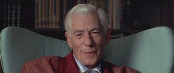 An older man sitting in a chair in front of a bookshelf