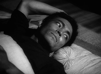 Movie still from “Godzilla” (1954), directed by Ishirô Honda – A black and white photo of a man laying in bed; Close Up shot, High angle
