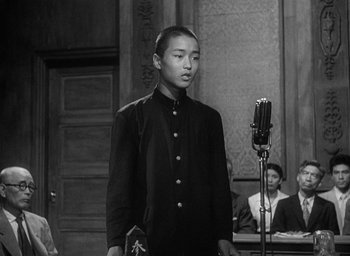 Movie still from “Godzilla” (1954), directed by Ishirô Honda – A man in a black suit standing in front of a microphone; Medium shot, Over the shoulder angle