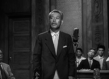 Movie still from “Godzilla” (1954), directed by Ishirô Honda – A man in a suit and tie standing in front of a microphone in front of other men; Medium shot, Low angle
