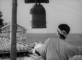Movie still from “Godzilla” (1954), directed by Ishirô Honda – A man standing next to a bell hanging from a wooden pole; Medium shot, Low angle