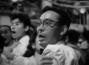 Movie still from “Godzilla” (1954), directed by Ishirô Honda – A man with glasses is singing into a microphone; Close Up shot, Low angle