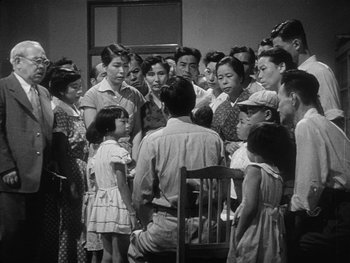 Movie still from “Godzilla: King of the Monsters!” (1956), directed by Ishirô Honda – A group of people gathered around a man in front of a mirror; Wide shot, High angle