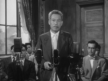 Movie still from “Godzilla: King of the Monsters!” (1956), directed by Ishirô Honda – A man in a suit and tie holding a microphone; Medium shot, Low angle