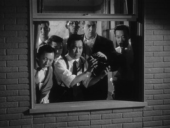 Movie still from “Godzilla: King of the Monsters!” (1956), directed by Ishirô Honda – Black and white photograph of a group of men looking through a window; Medium shot, Low angle