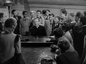 Movie still from “Going My Way” (1944), directed by Leo McCarey – A group of young men and women standing around a man playing a piano; Medium shot, High angle