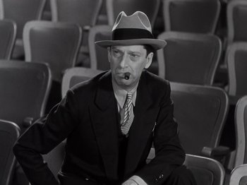 Movie still from “Gold Diggers of 1933” (1933), directed by Mervyn LeRoy – A man in a suit and a hat sitting in a row of seats; Medium shot, Low angle