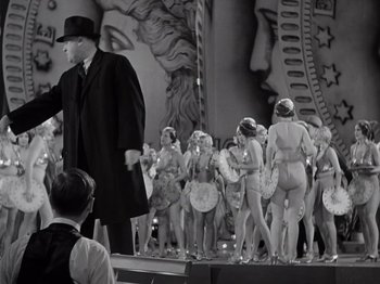Movie still from “Gold Diggers of 1933” (1933), directed by Mervyn LeRoy – A man in a black coat and hat standing in front of a group of naked women; Wide shot, Low angle