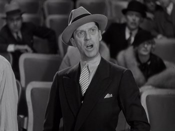 Movie still from “Gold Diggers of 1933” (1933), directed by Mervyn LeRoy – A man wearing a suit and a hat standing in front of a group of people; Close Up shot, Low angle