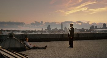Two men are sitting on the roof of a building and one is looking out at the city